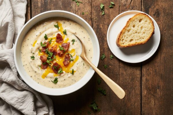 A steaming bowl of creamy potato soup garnished with chives, bacon, and a drizzle of cream, with a rustic bread roll on the side.