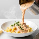 Creamy homemade Alfredo sauce being poured over fettuccine pasta in a white ceramic bowl