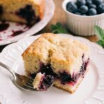 Slice of moist blueberry cake with a crackly sugar top, fresh blueberries, and a dollop of whipped cream on a vintage plate.
