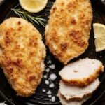 Golden-brown, perfectly fried chicken cutlets on a wire rack with lemon wedges and fresh herbs